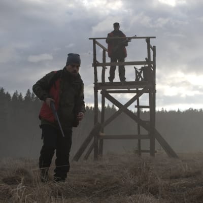 Joonas Saartamo med gevär i handen går mot kameran medan Peter Franzén håller utkik från ett jakttorn.