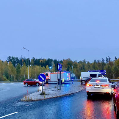En T-korsning i morgonskymning, våt asfalt, personbilar, en paket och en brandbil, trafikmärken som visar vägen till Karis, Hangö, Helsingfors och så vidare.. En situation där det har hänt en bilolycka.