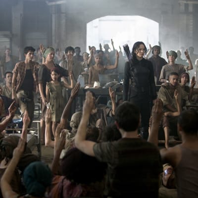 Katniss Everdeen står i en sal med en massa människor runt sig.