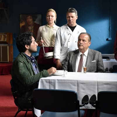 Khaled (Sherwan Haji) sitter vid ett bord och blir serverad soppa, vid bordet sitter även Wikström (Sakari Kuosmanen) och runtom står tre personer och ser Khaled.