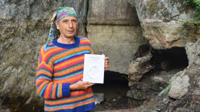 En medelålders man i randig tröja och duk om huvudet står framför en låg grottöppning. Han håller i en bok.