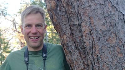 En medelålders man står mellan två trädstammar och ler. Den ena stamman, till höger, är en ovanligt stor tall.