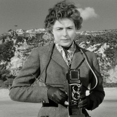 Tympääntyneen näköinen nainen (Ingrid Bergman) ottamassa valokuvaa Rolleiflex-kameralla rantamaisemassa. Kuva elokuvasta Matka Italiaan (Matka Italiassa, Viaggio in Italia).