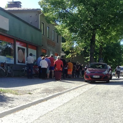 Folk köar utanför en tidigar mataffär där en förening delar ut mathjälp i Karis. Sommar. Bilar och cyklar syns också.