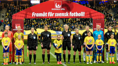 Lina står uppställd på rad på fotbollsplanen i en spelarena med juniorfotbollsspelare och de andra domarna. En stor röd skärm med texten "Framåt för svensk fotboll" står bakom dem.