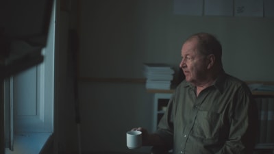 Roy Andersson sitter med en kaffekopp och tittar ut genom ett fönster.