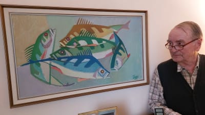En äldre man intill en tavla föreställande en abborre i rörelse. Mannen ser mot målningen där han står lutad emot en låg bokhylla. Han bär en rutig skjorta och en ärmlös stickad väst.