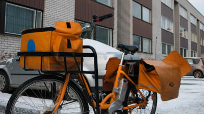 En postutdelares cykel med sina orange väskor parkerad framför ett höghus.