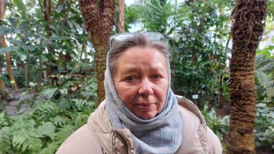 En kvinnlig författare som med en ljusblå duk på huvudet och en beige vinterkappa står sorgmodigt leende i Helsingfors Botaniska trädgård