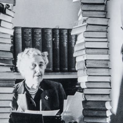 Agatha Christie katsoo kameraan kahden korkean kirjapinon välistä.