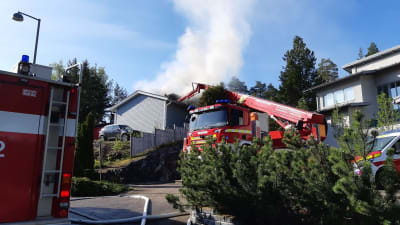 En brandbil står på en gårdsplan. I bakgrunden syns vit rök stiga upp från ett hus. 
