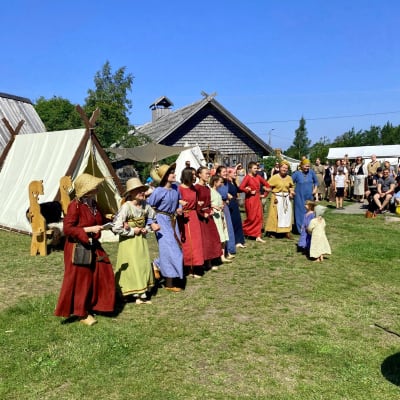 Många människor i vikingakläder dansar i ett långt led på en gräsmatta framför hus byggda i vikingastil.