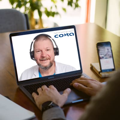 Pöydällä on tietokone, jossa on auki videoyhteys lääkäriin, joka hymyilee kuulokkeet korvilla ja mikrofoni suun edessä.