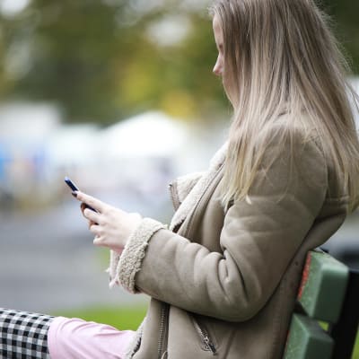 Ung kvinna sitter på en bänk med mobiltelefon i handen, 