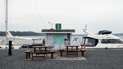 Två bord med bänkar står ute vid en pir. I bild synd två båtar och en rätt stor informationstavla för turister.