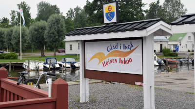 En skylt där det står Glad i Ingå på svenska och finska. I bakgrunden syns en småbåtshamn.
