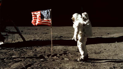 Vi är uppe i rymden. En man är klädd i en helvit rymddräkt. Han vandrar på en plan sandyta. I mitten på planen står den amerikanska flaggan. På sandytan syns märken av mannens skor. Bakgrunden är svart.