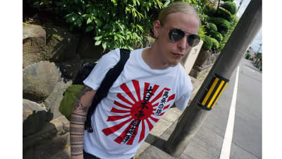 En man med långt blont hår. Han bär solglasögon och en t-shirt med med japansk text på.