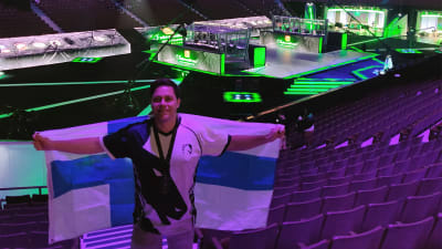 En man står inne i en stor arena. Han håller i en stor Finlands flagga. Ljuset på honom är lila. I bakgrunden syns stora skärmar och ett område för dataspelare. 