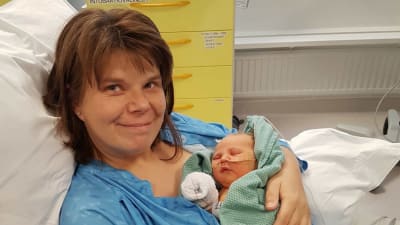 Mamma med nyfött spädbarn i armarna. De ligger på en sjukhusbädd. Mamman ler och bebisen sover.