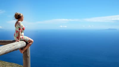 Kvinna i blommig klänning som sitter på ett staket och blickar ut över ett blått hav. Också himlen är blå.