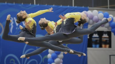 Tre unga kvinnor i gul-grå gymnastikdräkter gör spagat i luften