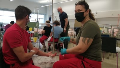 En kvinna tar blodprov på en man. Det här sker i undervisningssyfte i ett stort rum med många studerande som gör samma sak.