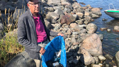 En äldre man, Folke Österman står på en stenig strand. I hans händer är en stor kasse med plastskräp.