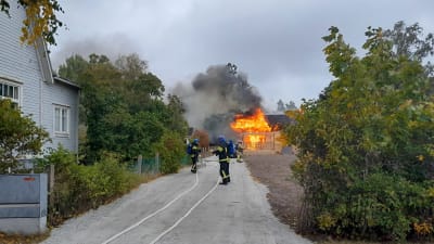 Ett hus brinner i bakgrunden, brandmän på väg att släcka