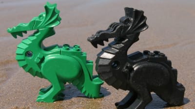 Två legodrakar, en grön och en svart, på en sandstrand.