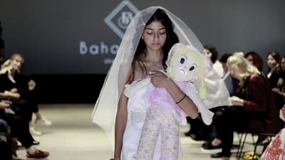Rauan Badawieh en klänning designad av Bahaulddin Rawi. Modellen går på catwalken och håller en docka i famnen. 