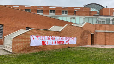 En tegelbyggnad med en banderoll där det står "Vinn den viktigast matchen, vi hejar på er" på italienska.