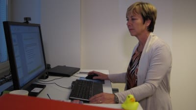 Kvinna i kort hår står vid sitt arbetsbord och tittar på dataskärmen