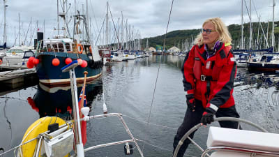 Bettina står i seglarkläder i aktern på en segelbåt som står i hamnen. Hon håller i rodret och ser mot vänster.