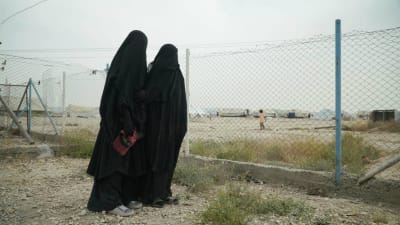 Två kvinnor iklädda heltäckande niqabs står vid ett nätstängsel på IS-lägret. En av kvinnorna har en röd keps i handen.