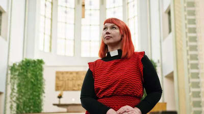 Pastor Marjaana Toiviainen i röd klänning. Bilden är tagen lätt underifrån och Marjaana har blicken vänd uppåt.