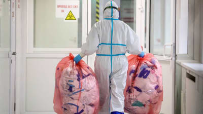 En person med skyddsutrustning bär två stora plastpåsar med avfall i händerna. Personen går mot en dörr i en sjukhuskorridor.