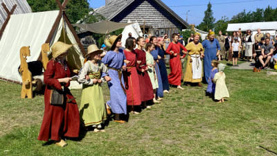 Många människor i vikingakläder dansar i ett långt led på en gräsmatta framför hus byggda i vikingastil.