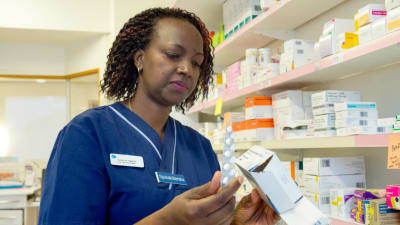 En sjuksköterska i klädd arbetsuniform står och inspekterar en pillerkarta i sin hand. I andra handen har hon en förpackning med mediciner.