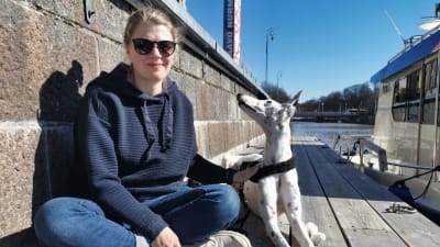 En ung kvinna med solglasögon sitter på en brygga med en hund bredvid sig som tittar på henne. 