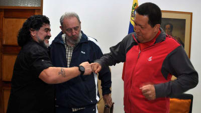Diego Maradona och Venezuelas tidigare ledare Hugo Chavez hälsar på varandra. Båda skrattar.  I bakgrunden står Kubas tidigare ledare Fidel Castro