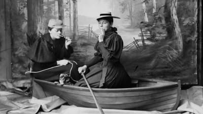 Marie Høeg och Bolette Berg i ett ateljéearrangemang där de klädda i kvinnokläder sitter rökande i en eka i slutet av 1800-talet.