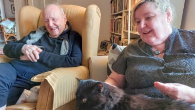 Författaren och diplomaten Lars Bergquist skrattar i sin länstol med författaren och historikern Carina Nynäs sittande bredvid sig i soffan. Också hon ser glad ut med katten Felix i famnen. November 2021.