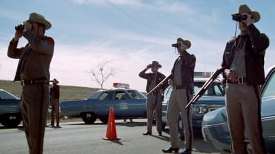Poliisin tiesulku amerikkalaisella maantiellä, joukko haulikoin aseistautuneita poliisimiehiä seisoo autojensa luona tähystämässä kiikareilla.