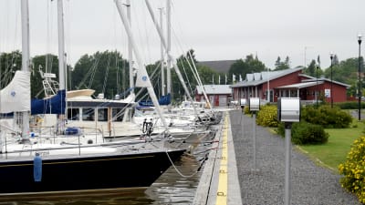 Flera båtar står förtöjda vid en brygga i en hamn. I bakgrunden syns ett rött hus.