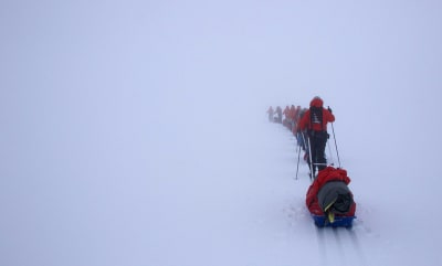 Sju personer skidar på rad. Alla släpar på pulkor bakom sig. Det är dimmigt och man ser inte horisonten. Bara snö. 