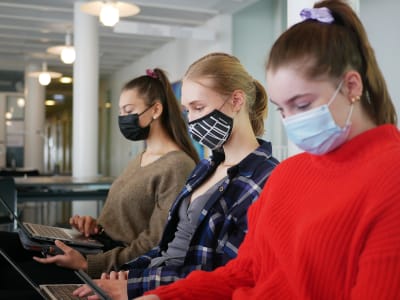 Studerande som har munskydd på sig och som sitter framför sina bärbara datorer vid en soffa i Svenska social- och kommunalhögskolans utrymmen i Kronohagen i Helsingfors.