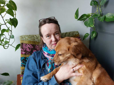 En leende kvinna sitter i sitt arbetsrum med sin hund i sin famn.