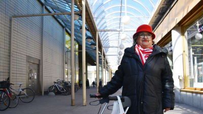 En äldre dam i svart jacka och röd hatt står bredvid en rollator. i stadsmiljö. Hon ler och tittar snett uppåt.