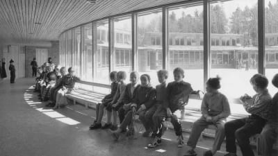 Koululaisia istuu koulun käytävällä, joka kaareutuu ja jonka seinä heidän takanaan on kokonaan lasia. Kuva Carl Nyrénin suunnittelemasta Danderydin Balderskolanista.